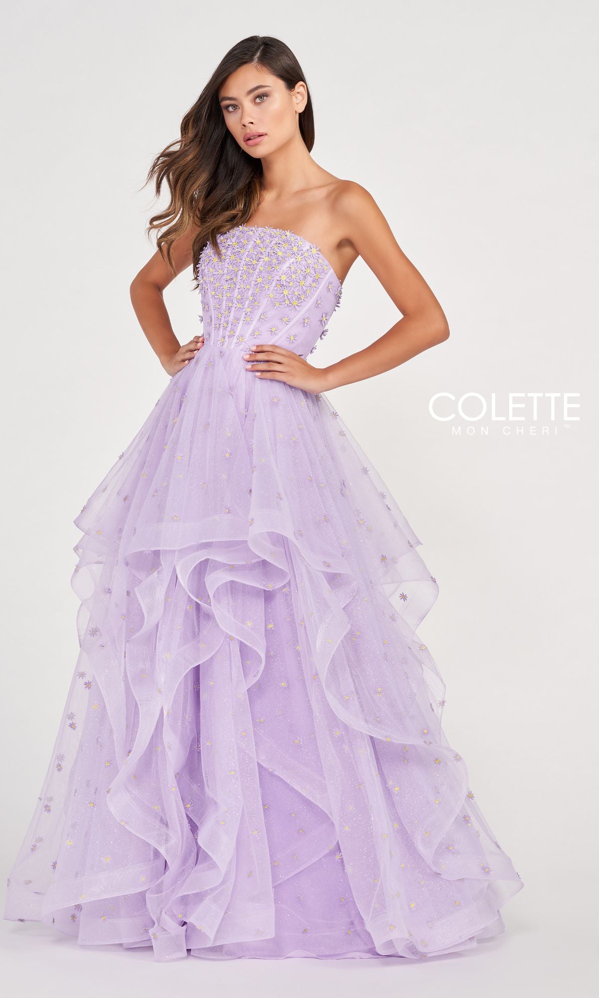 A woman poses with her hands on her hips in the Colette CL2055 Strapless Daisy Ball Gown, a floor-length lavender tulle dress featuring layered skirts and decorative beadwork, against a plain background.